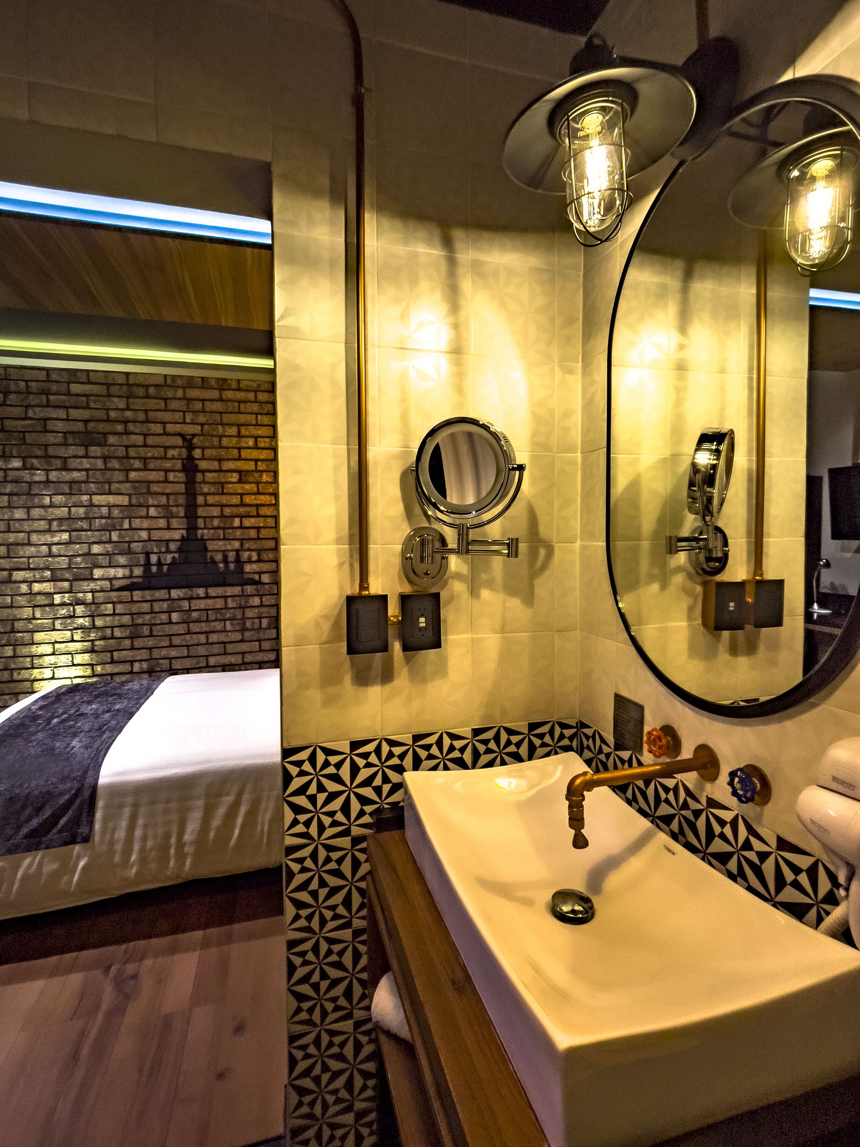 A hotel bathroom with a sink, mirror, and towel rack. There is a patterned wall and a bed in the background.
