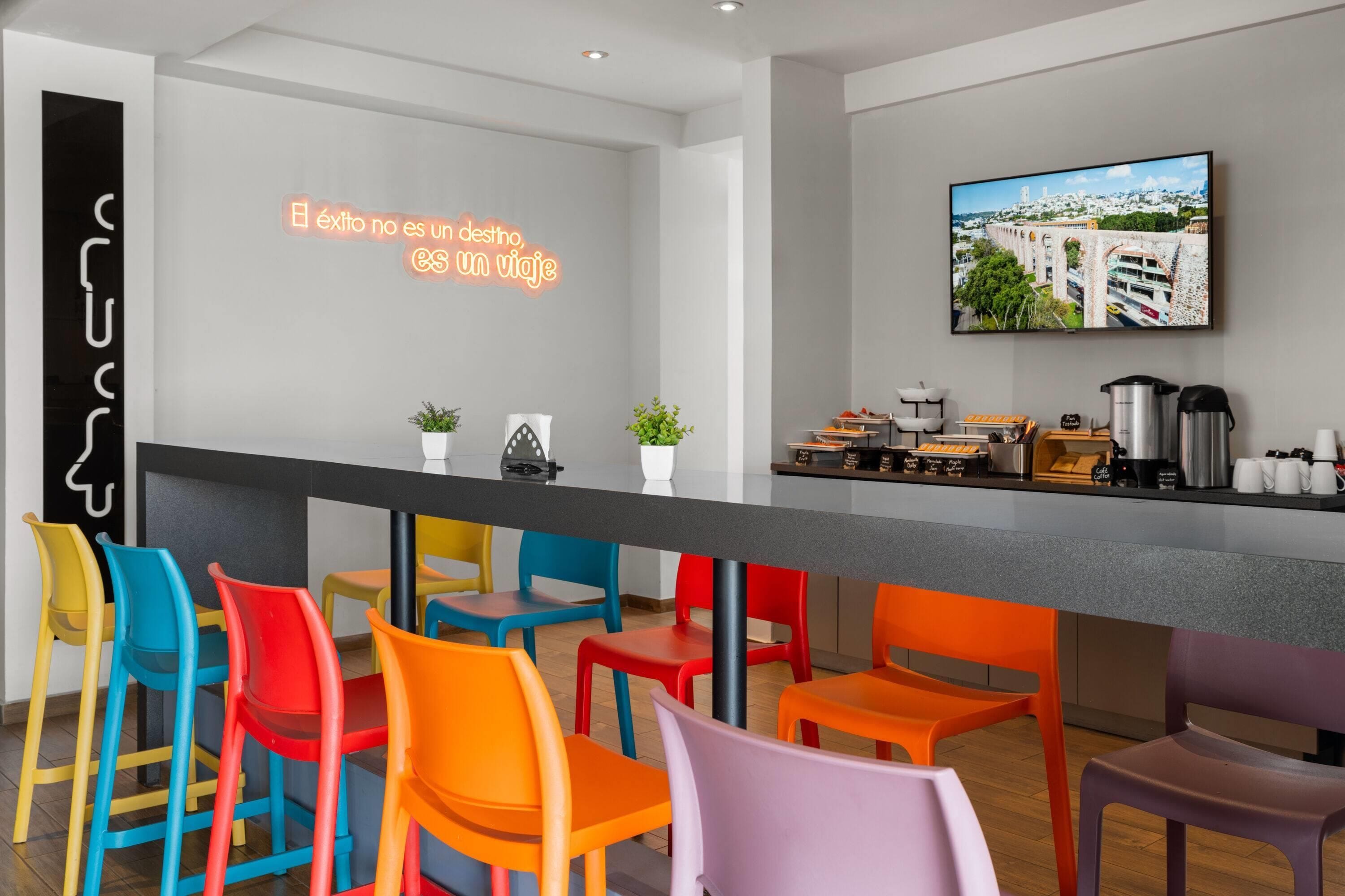 Een moderne bar met kleurrijke stoelen, een neonbord, een koffiehoek en een televisiescherm met stadsgezicht.