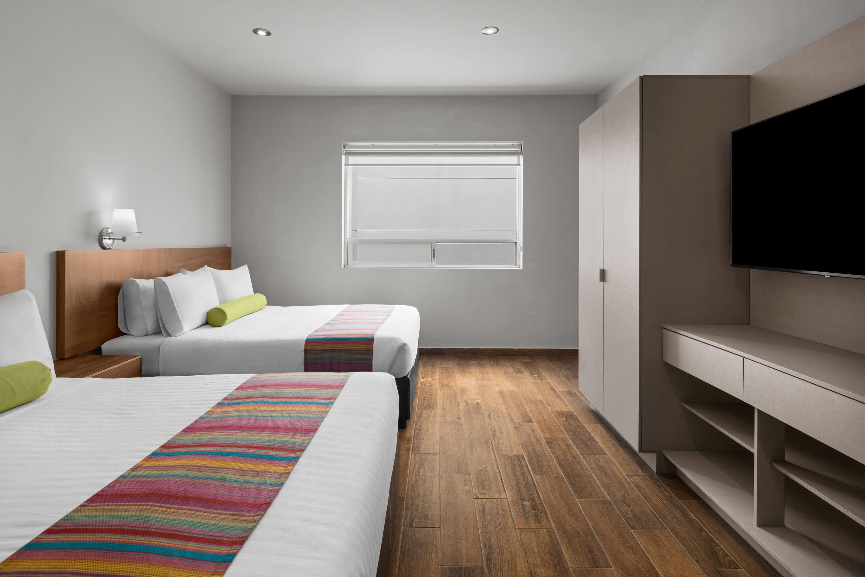 A modern hotel room with two beds, a television, and a window with blinds.