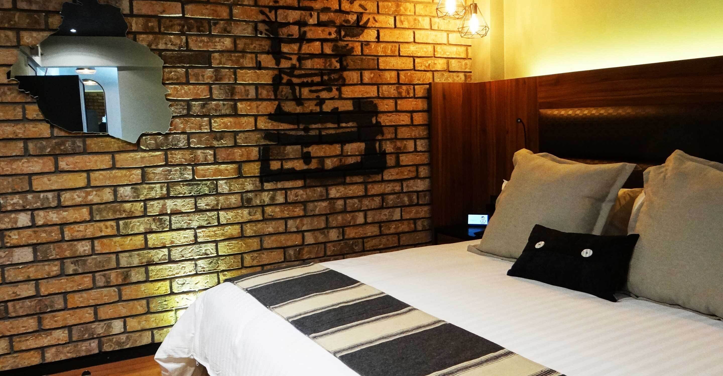 A hotel room with a brick wall, a bed with striped bedding, and a nightstand with a lamp.