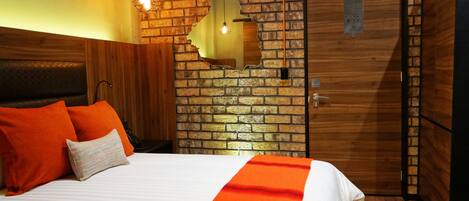 Habitación de hotel con pared de ladrillo, puerta de madera, cama con ropa blanca y naranja, y dos lámparas colgantes.