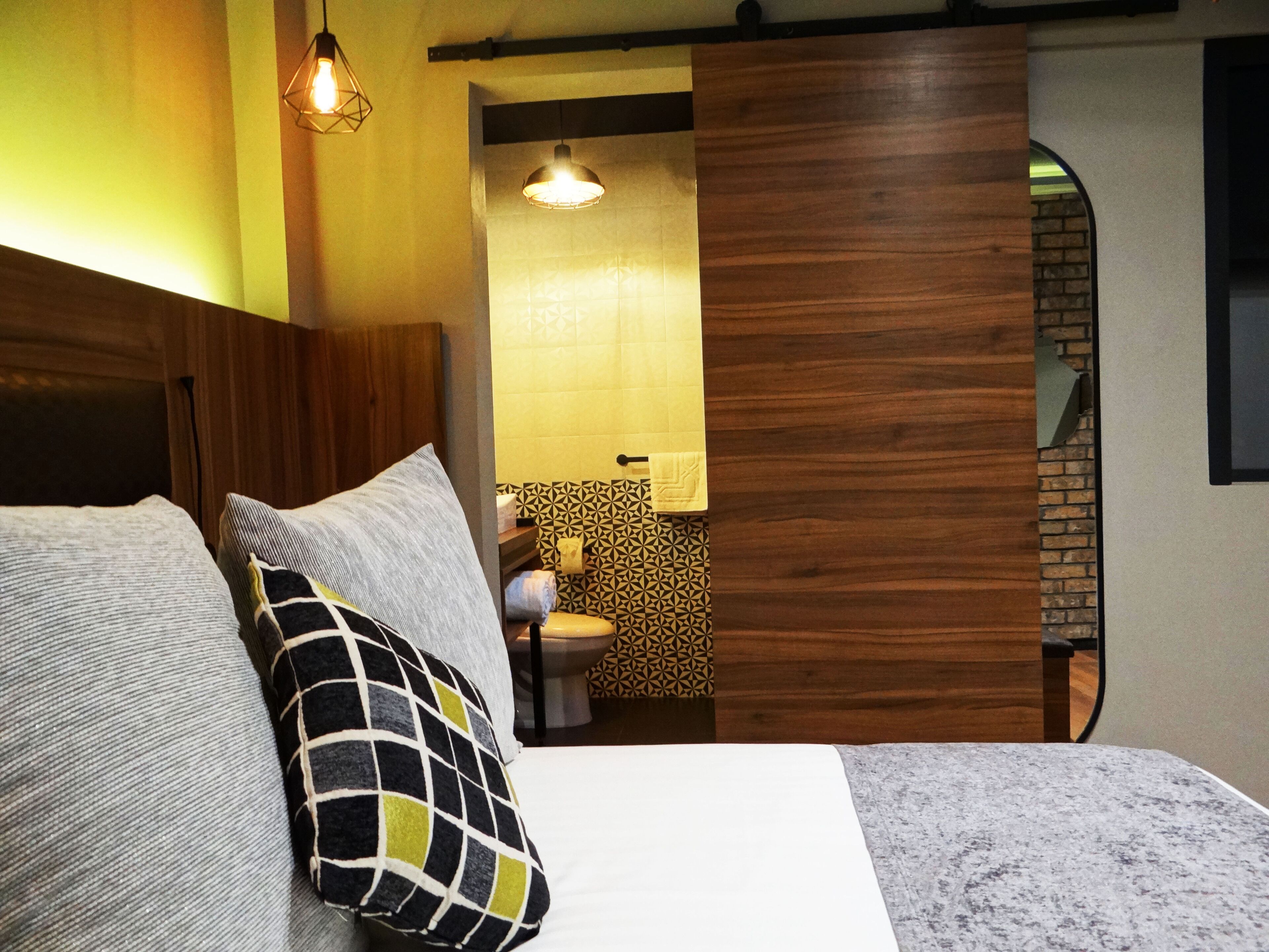 Een slaapkamer met een bed, kussens, een houten hoofdbord, een hanglamp en een badkamer die door een open deur zichtbaar is.