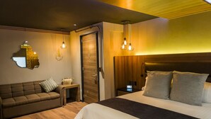 A modern bedroom with a wooden headboard, a large bed with grey bedding, a brown sofa with striped pillows, a small bedside table with a lamp, and a mirror on the wall. - Cadillac Hotel Boutique (Mexico City)