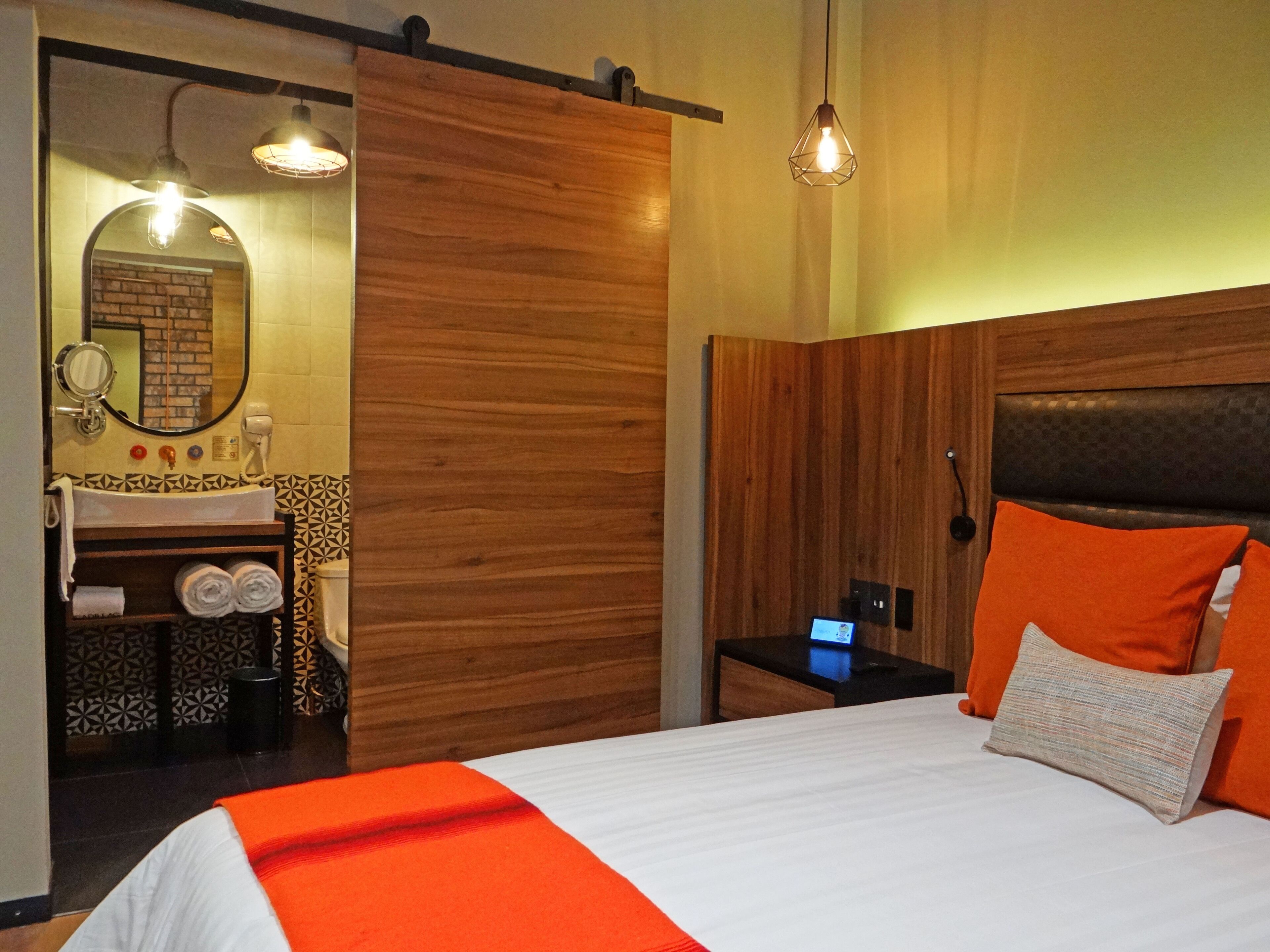 A hotel room with a wooden headboard, a bed with an orange throw, a nightstand with a lamp, and a bathroom with a mirror, sink, and towel rack.