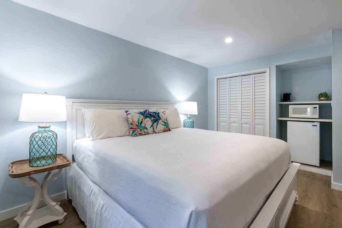 A bedroom with a bed, two bedside tables with lamps, a wardrobe, and a small kitchenette.