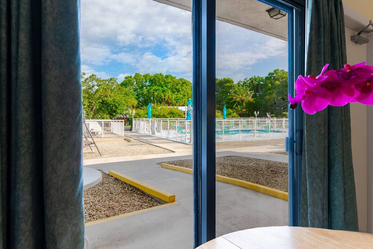 A view through a window of a concrete pathway leading to a fenced area with green chairs and trees in the background.