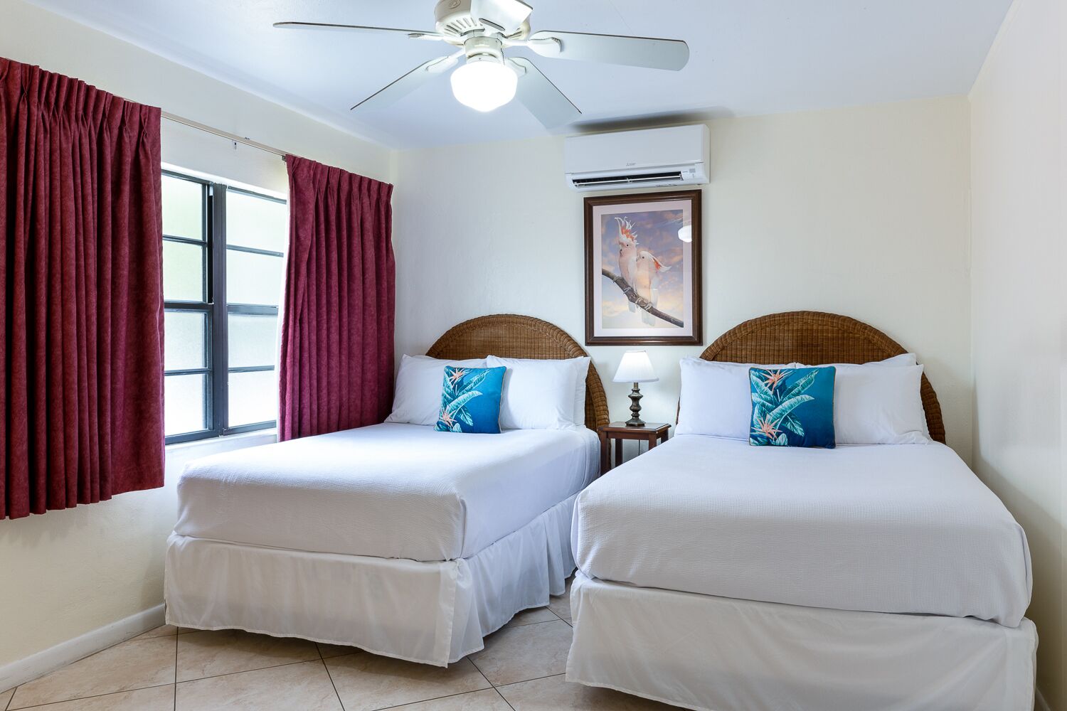 Une chambre d’hôtel avec deux lits simples, un ventilateur de plafond, un appareil de climatisation et une fenêtre avec des rideaux rouges.