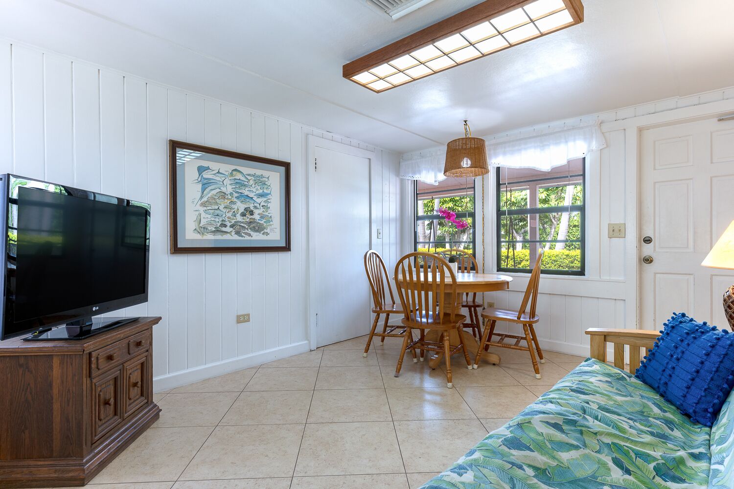 A room with a TV on a wooden cabinet, a dining table with chairs, a sofa with a leaf-patterned cover, and a window with a view of greenery.