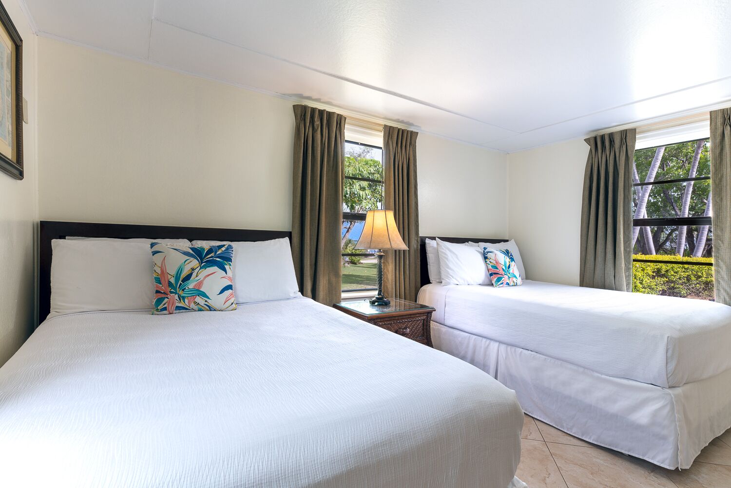 Dos camas individuales con ropa de cama blanca y almohadas con motivos tropicales, una mesita de noche con lámpara, y vistas a la vegetación a través de una ventana con cortinas.