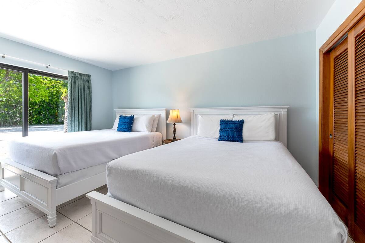 A hotel room with two beds, a window with curtains, a lamp, and a view of greenery outside.