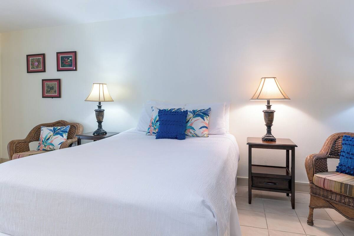 A hotel room with a large bed, two nightstands with lamps, wicker chairs, and framed pictures on the wall.
