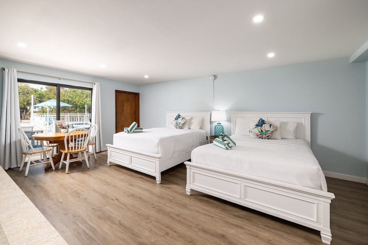 A hotel room with two beds, a dining area with a table and chairs, and a view of a pool and greenery outside.