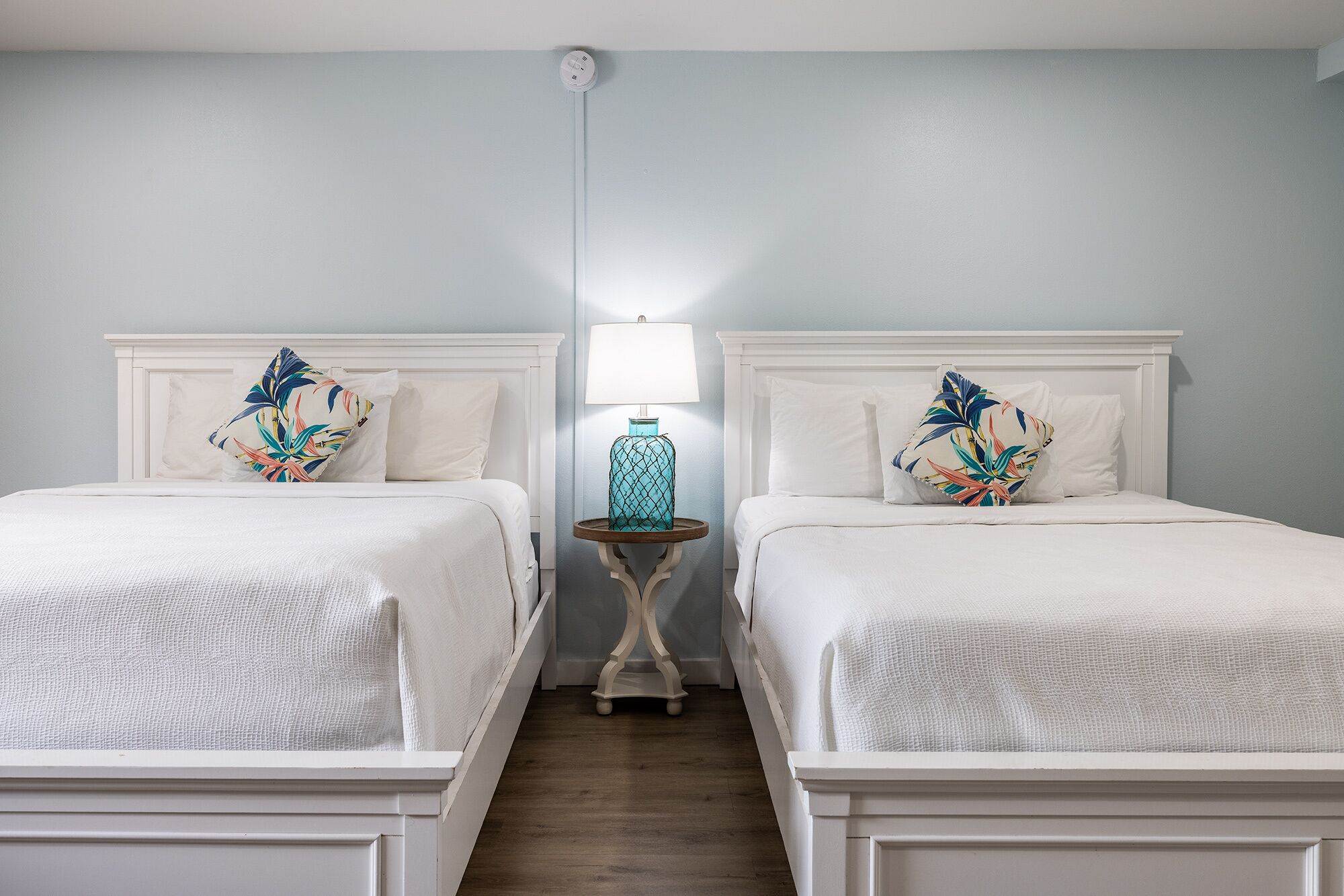 Une chambre d’hôtel avec deux lits, chacun doté d’une tête de lit blanche et d’un coussin à motifs floraux. Une petite table ronde avec une lampe se trouve entre les deux lits.
