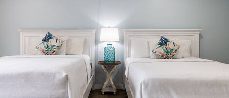 Une chambre d’hôtel avec deux lits, chacun doté d’une tête de lit blanche et d’un coussin à motifs floraux. Une petite table ronde avec une lampe se trouve entre les deux lits.