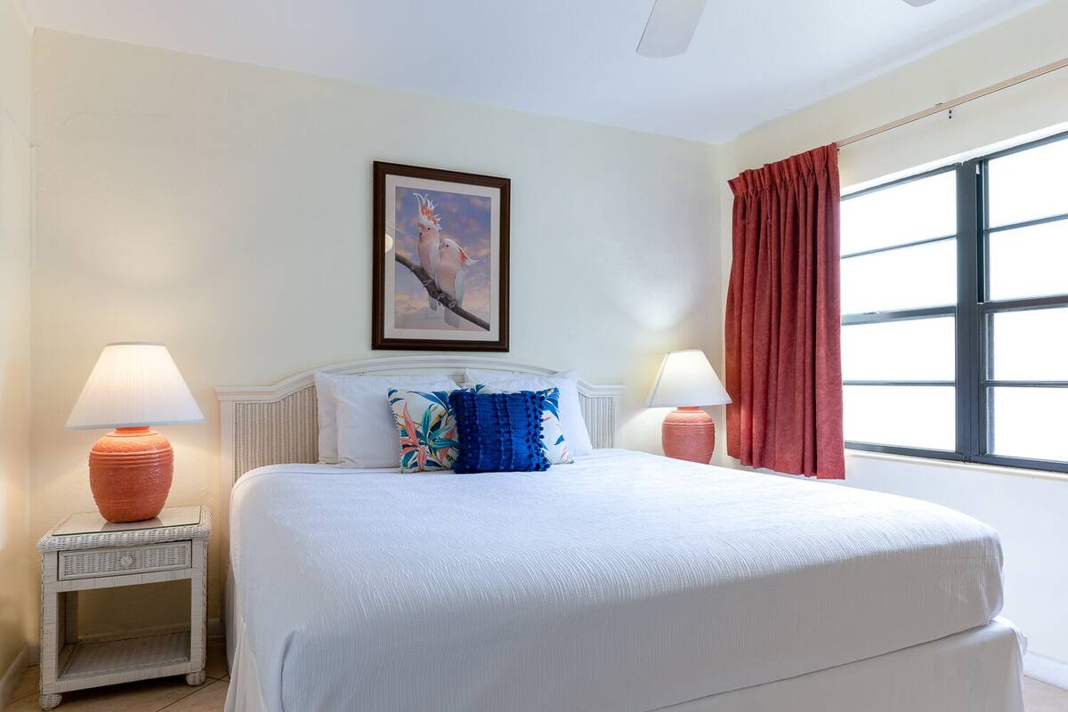 A hotel room with a large bed, two bedside tables with lamps, a framed picture on the wall, and a window with red curtains.