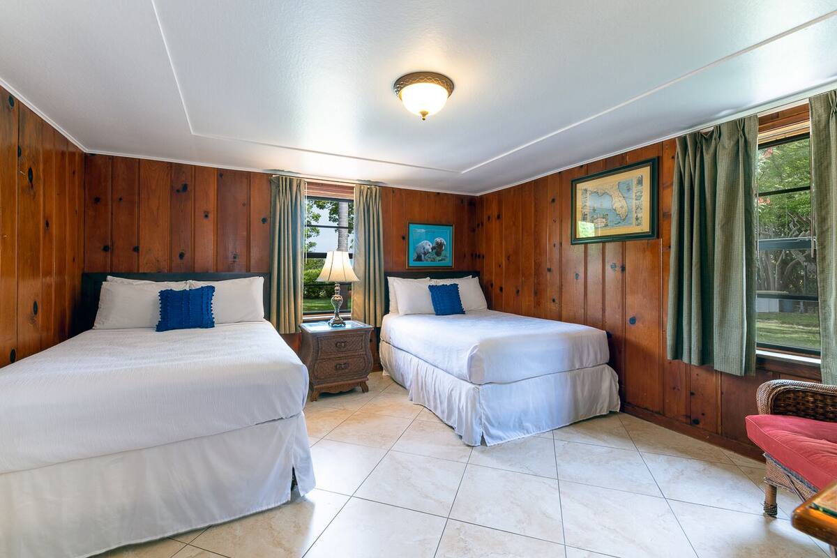 A room with two beds, wooden walls, a tiled floor, a nightstand with a lamp, and a framed picture on the wall.