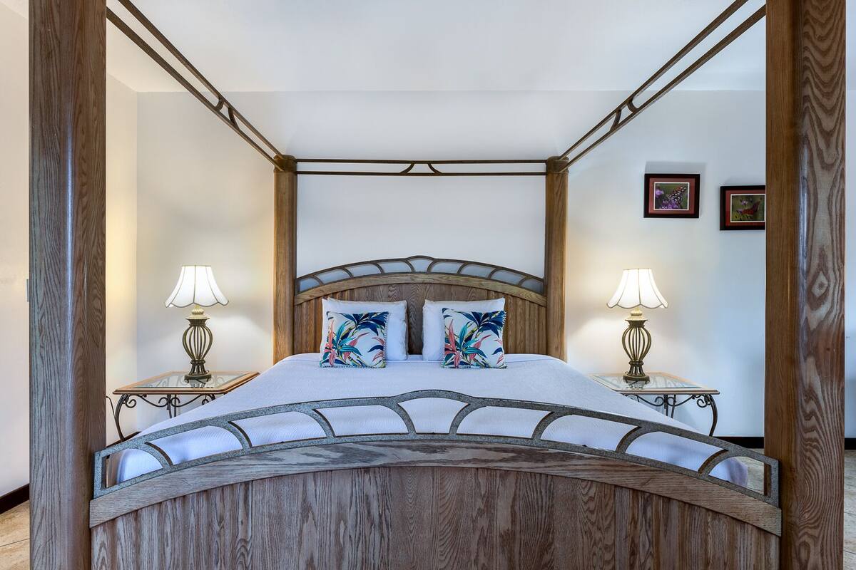 A four-poster bed with a wooden frame and a canopy, two bedside lamps, and two pillows with tropical prints.