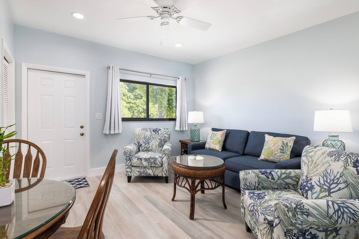 A living room with a blue sofa, two patterned armchairs, a glass coffee table, and a dining table with chairs.