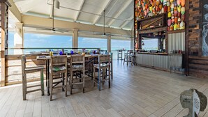 Un espace intérieur spacieux avec une table et des chaises en bois, un coin bar avec des bouteilles aux couleurs vives, et une vue sur l’océan.