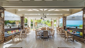 Un vaste espace extérieur aménagé pour les repas, avec des tables et des chaises en bois, une table ronde centrale et une vue sur l’océan.