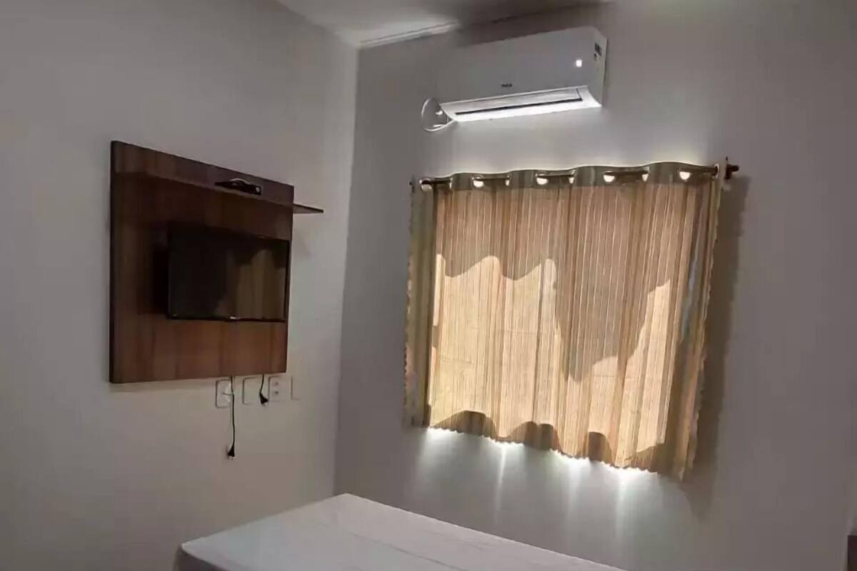 A single-bed room with a flat-screen TV mounted on the wall, a window with sheer curtains, and a ceiling air conditioner.