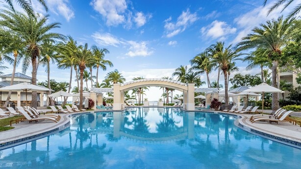 A large pool with lounge chairs and umbrellas, surrounded by palm trees and a clear sky.