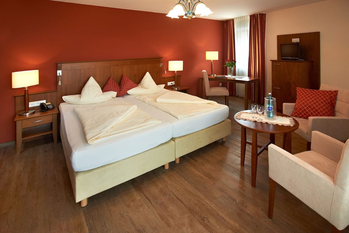 Ein Hotelzimmer mit Doppelbett, Nachttischen mit Lampen, einem Schreibtisch mit Stuhl, einem Fernseher auf einer Kommode und einem kleinen runden Tisch mit Flasche und Gläsern.