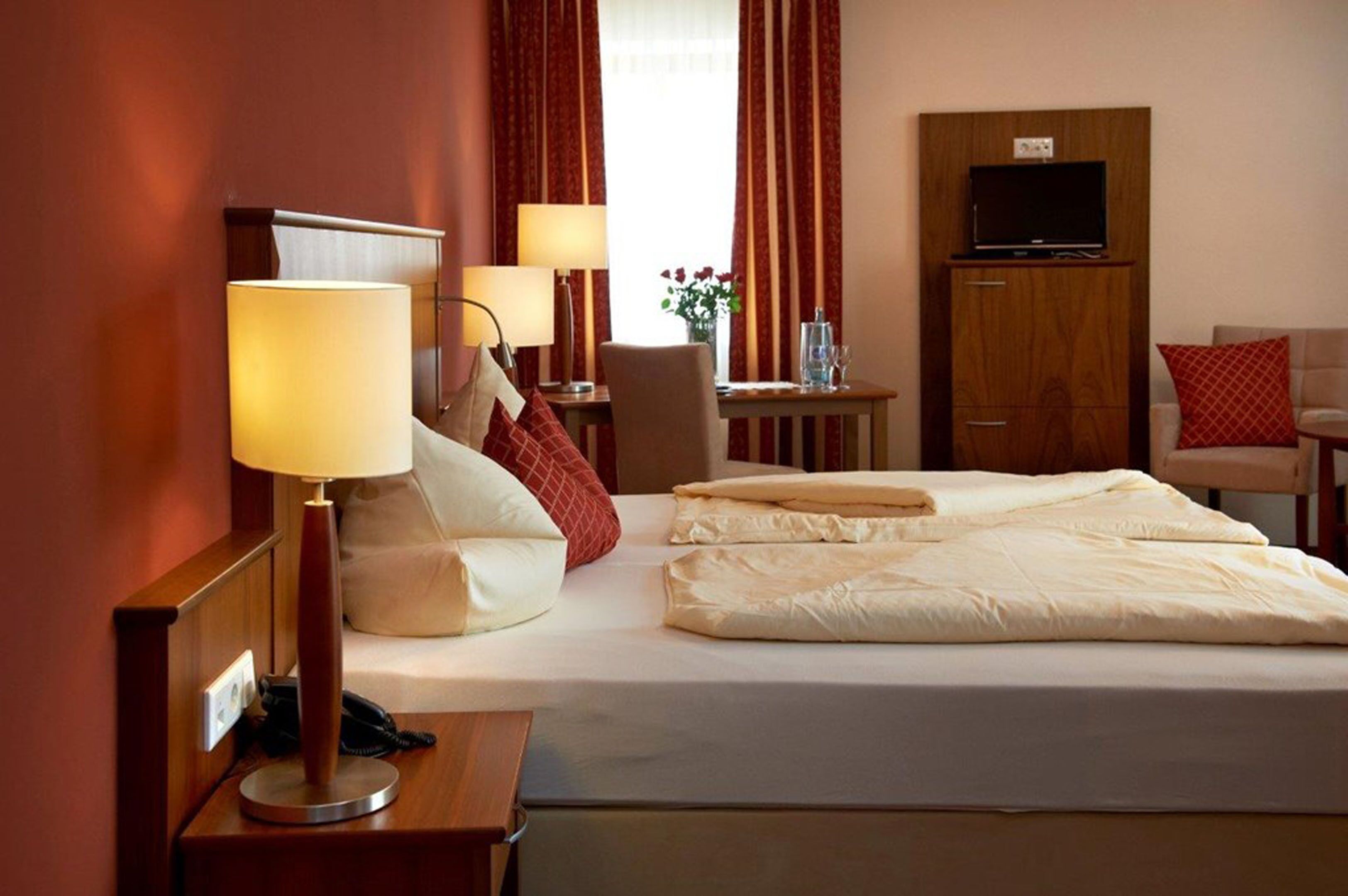 Habitación de hotel con cama, lámparas de noche, escritorio con silla, televisor sobre un mueble de madera y ventana con cortinas.