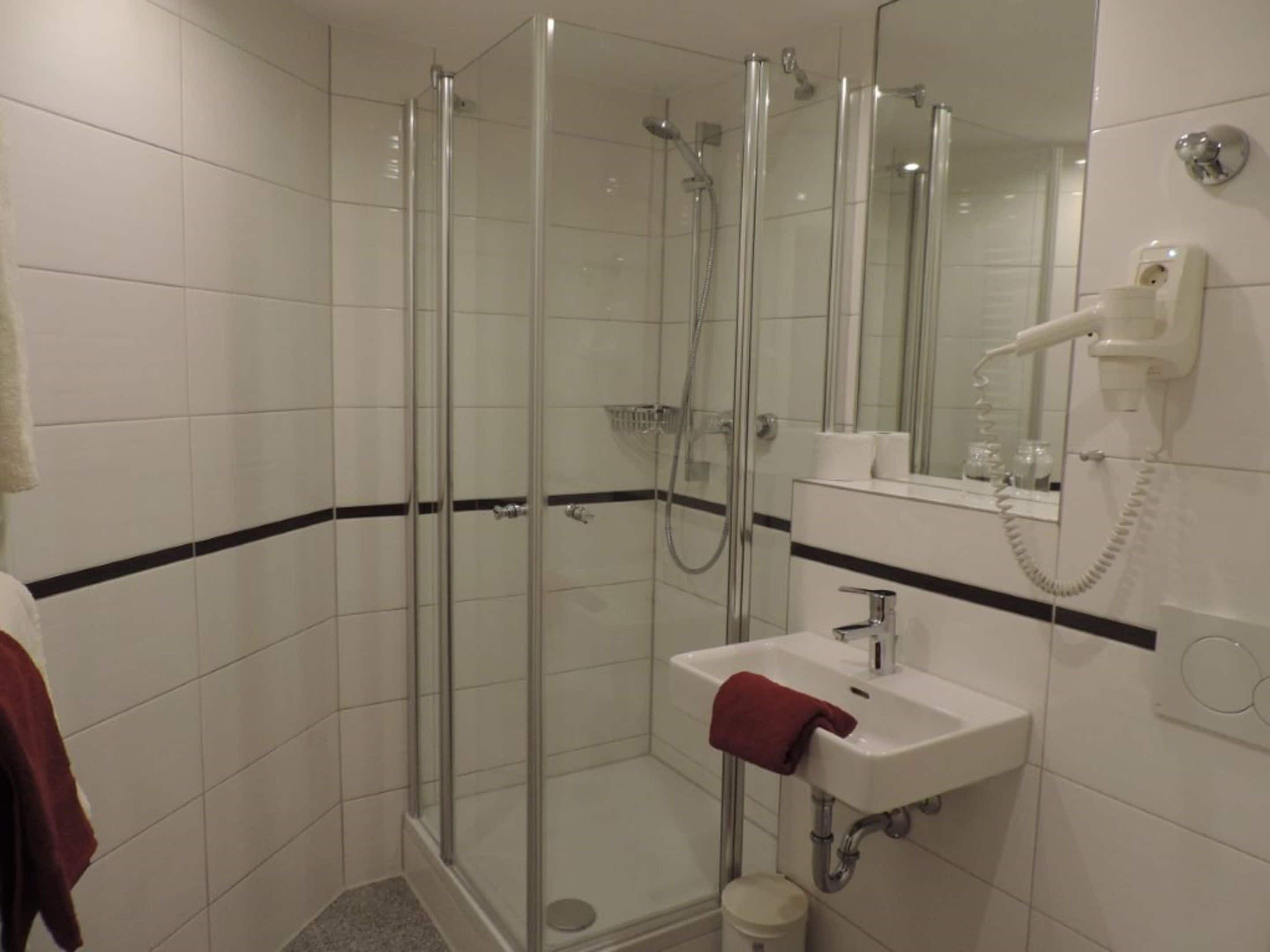 Un box de ducha con paredes de vidrio, un grifo de ducha de mano, un lavabo blanco con una toalla roja y un secador de pelo en un soporte fijado a la pared.