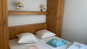 A wooden bed with two pillows, a folded towel, and a blanket. There are two wall-mounted lamps on a shelf above the bed.