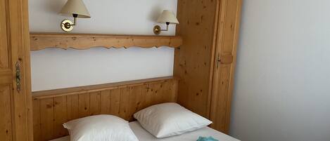 En træseng med to puder, et foldet håndklæde og et tæppe. Der er to vægmonterede lamper på en hylde over sengen.