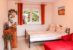 A bedroom with a bed, a red sofa, a wooden dresser, a window with curtains, and a painting on the wall.