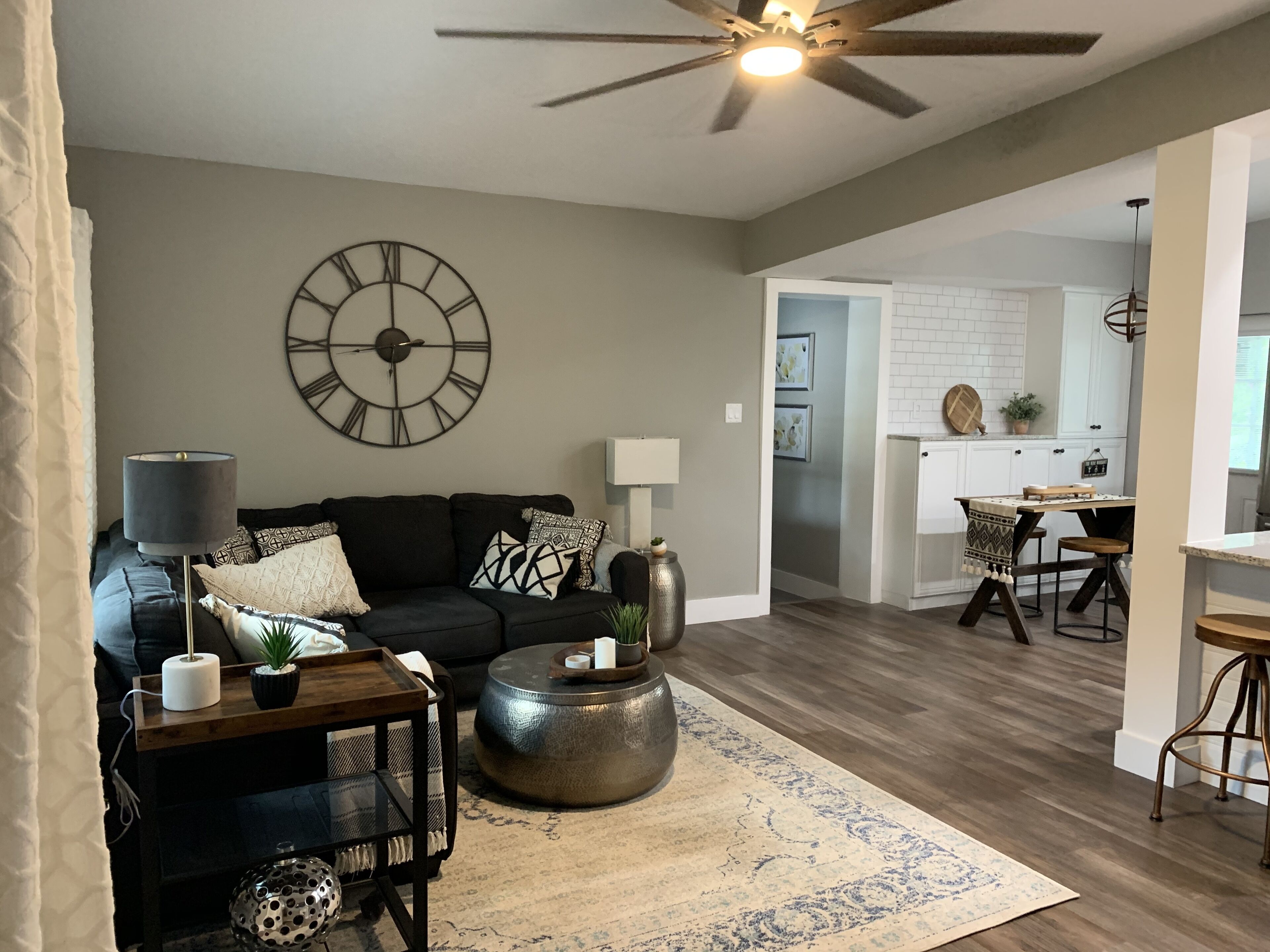 A living room with a black sofa, a round coffee table, and a large wall clock. There is a dining area with a table and chairs visible in the background.