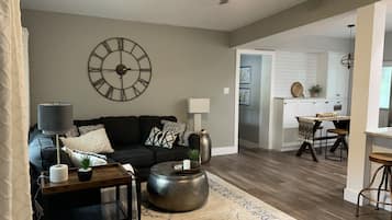 A living room with a black sofa, a round coffee table, and a large wall clock. There is a dining area with a table and chairs visible in the background.