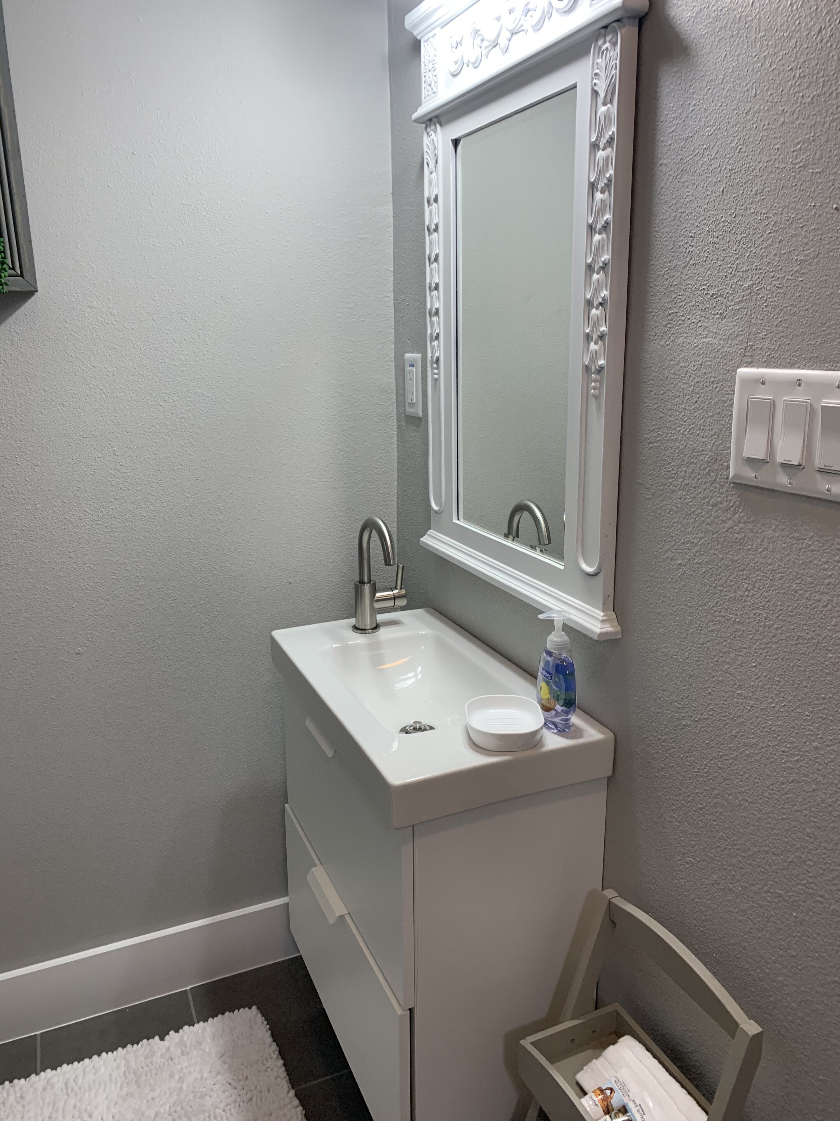 A bathroom with a white sink, a mirror with decorative molding, a white towel dispenser, and a small storage box.