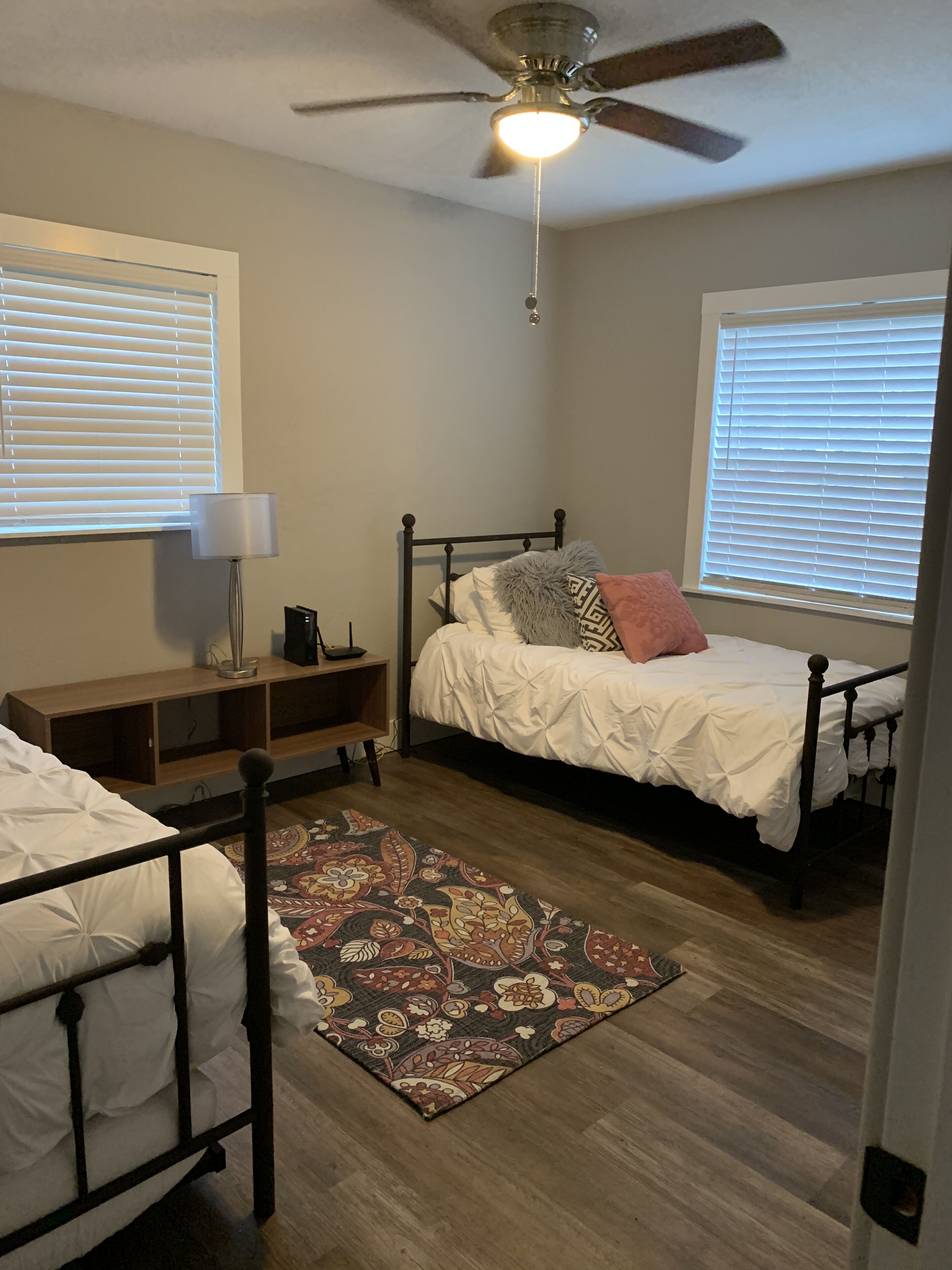A bedroom with two single beds, a ceiling fan, a nightstand, a lamp, and a patterned rug.