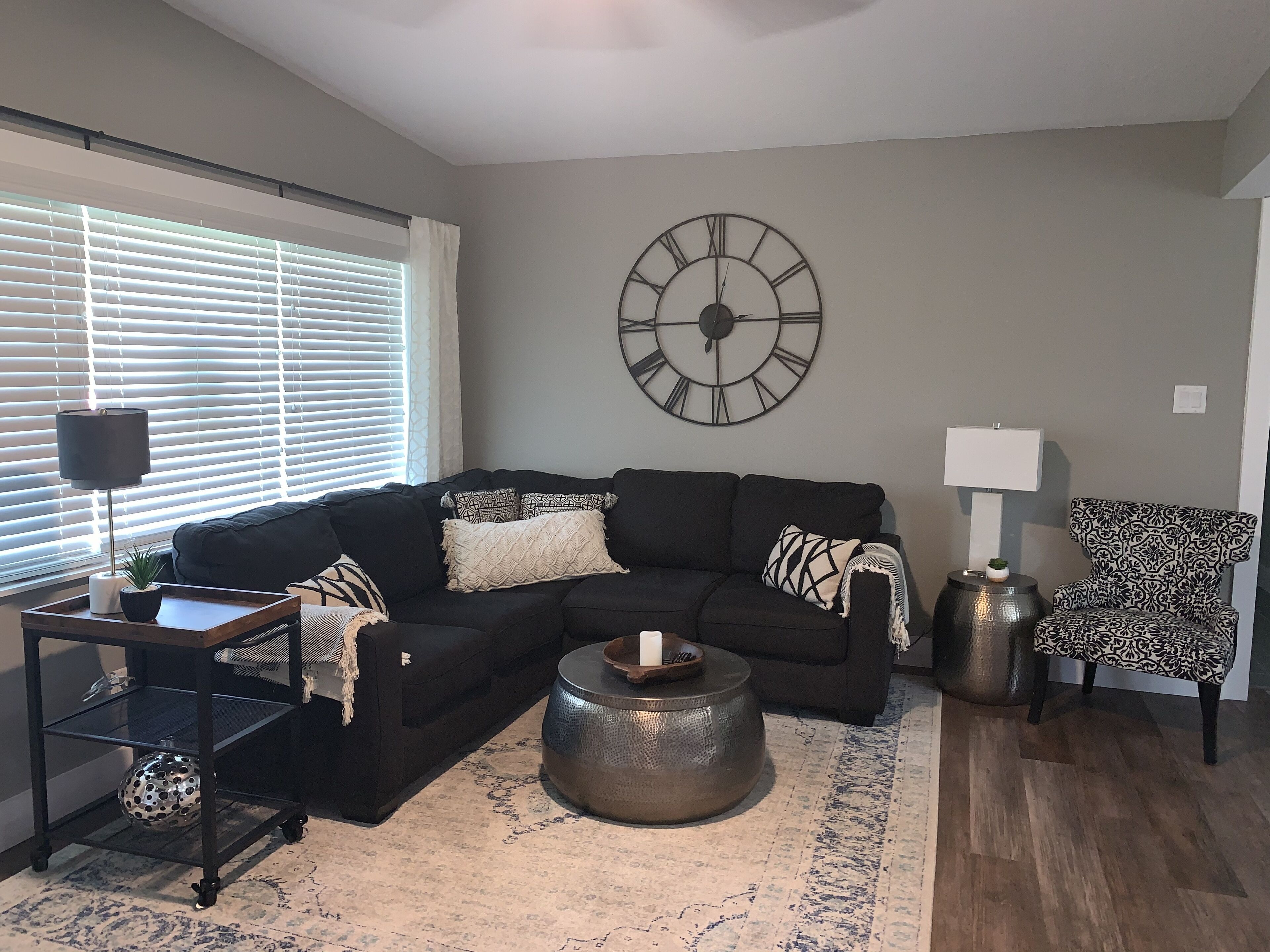 A living room with a sectional sofa, a round coffee table, a patterned armchair, a side table with a lamp, and a wall clock.