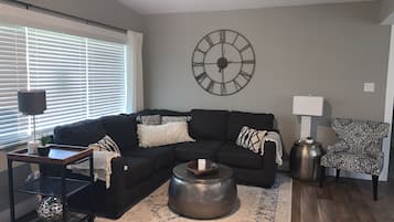 A living room with a sectional sofa, a round coffee table, a patterned armchair, a side table with a lamp, and a wall clock.