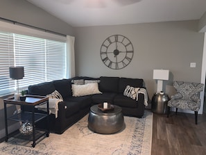 A living room with a sectional sofa, a round coffee table, a patterned armchair, a side table with a lamp, and a wall clock. - Mid Century Gem Near Theme Parks (Ocoee)