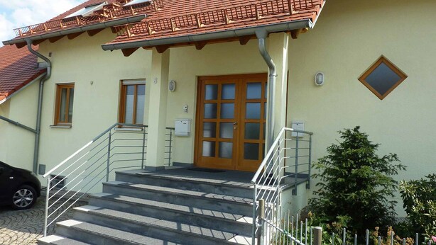 A building with a red-tiled roof, a wooden door, and a set of stairs leading to a porch with a metal railing. - Ferienwohnung Fantaisie (Eckersdorf)