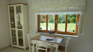 A dining area with a wooden table and white chairs, a glass cabinet with dishes, and a window with a view of greenery. - Ferienwohnung Fantaisie (Eckersdorf)