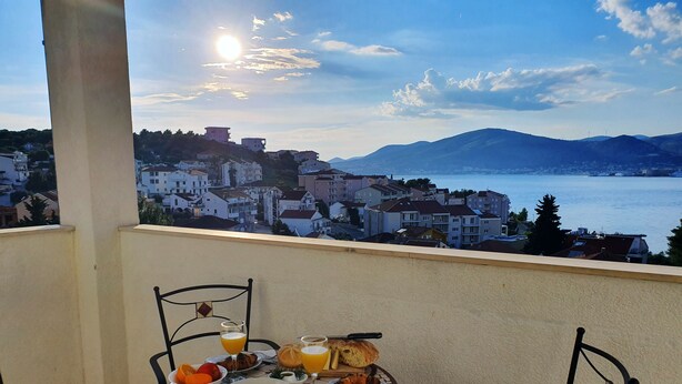 Varanda com mesa posta para o café da manhã, com vista para uma cidade costeira e montanhas.