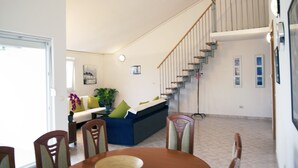 Uma área de refeições com mesa e cadeiras de madeira, uma escada que leva a um andar superior, e vista para a sala de estar ao fundo.