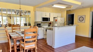 A modern kitchen with white cabinetry, a central island, and a dining area with wooden chairs and a table. - Nice apartment with golf course view in prestigious Brunswick Golf. Fits 4.  (Calabash)