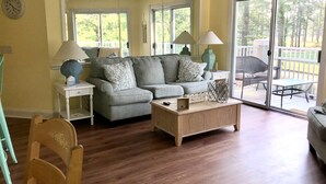 A living room with a grey sofa, wooden coffee table, and a sliding glass door leading to an outdoor deck. - Nice apartment with golf course view in prestigious Brunswick Golf. Fits 4.  (Calabash)