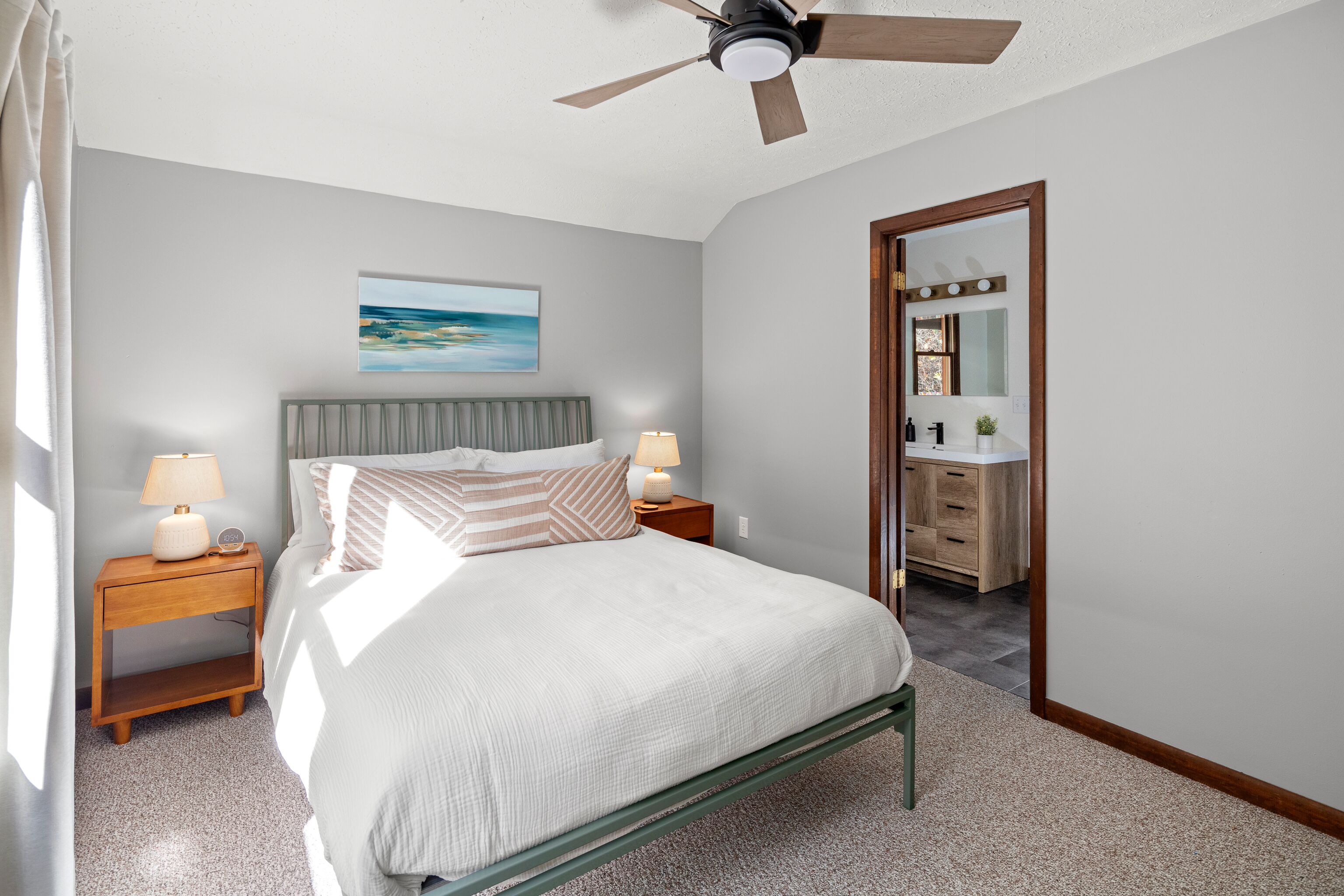A bedroom with a bed, nightstands, a ceiling fan, a painting, and a bathroom visible through an open door.