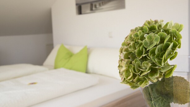 A neatly made bed with white bedding and a green pillow, a green plant in a glass vase, and a wall-mounted picture frame. - Pension Jederzeit (Wallersdorf)