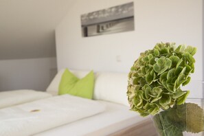 Una cama bien hecha con ropa blanca y una almohada verde, una planta verde en un florero de vidrio, y un marco de pared.