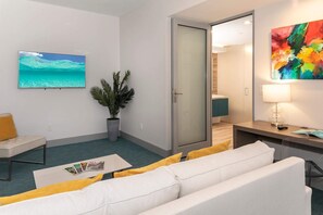 Ein modernes Hotelzimmer mit einem großen Flachbildfernseher, einem Sofa, einem Couchtisch, einer Topfpflanze, einem Schreibtisch mit einer Lampe und einem farbenfrohen abstrakten Gemälde.