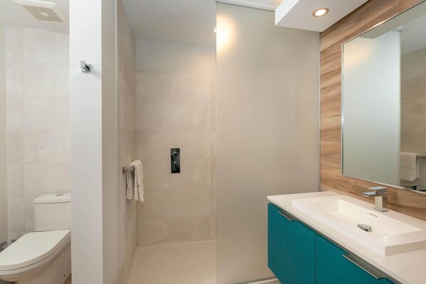 A modern bathroom with a white toilet, a glass shower enclosure, a white sink with a wooden vanity, and a large mirror. - Suite 11 with Large Bathroom (Cruz Bay)